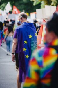Im Zentrum des Fotos ist eine männlich wirkende Person, die man von hinten sieht. Sie trägt einen Regenbogenmundschutz und als Cap eine Europa-Flagge, die in eine Regenbogenflagge übergeht. Wobei vor allem die EU-Flagge zu sehen ist. Denn der Mensch wird teilweise von einer unscharfen Person im rechten Bildvordergrund verdeckt, die ein regenbogenfarbenes Oderteil trägt. Im Hintergrund sind weitere Menschen zu sehen. Das Bild ist offenbar auf einer Pride-Demo entstanden.